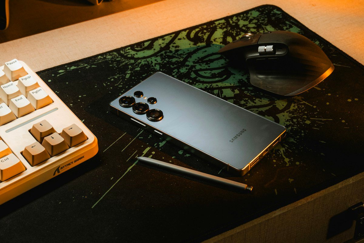A smartphone lies beside a keyboard and mouse in warm window light.